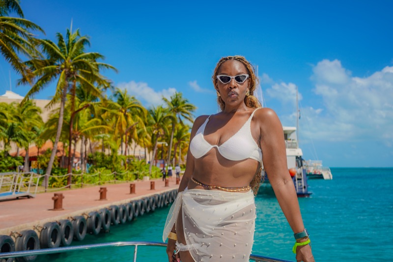 Woman in a white swim set and cat-eye sunglasses standing on a boat rail against turquoise water.