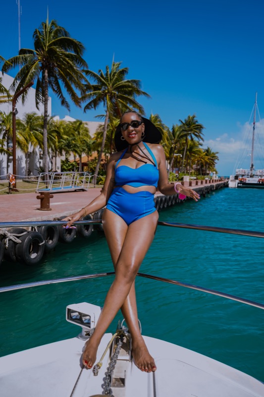 Woman in a blue two-piece and wide sun hat perched on a yacht bow, marina behind.