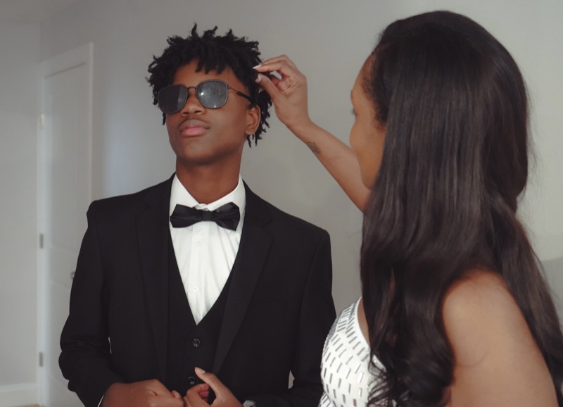 Mother in a white beaded dress adjusting her son's hair as he stands in a black tuxedo and sunglasses.