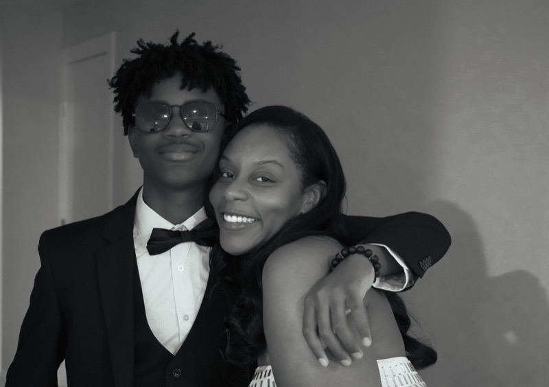 Black-and-white portrait of a smiling mother wrapped in her tuxedoed son's arm.