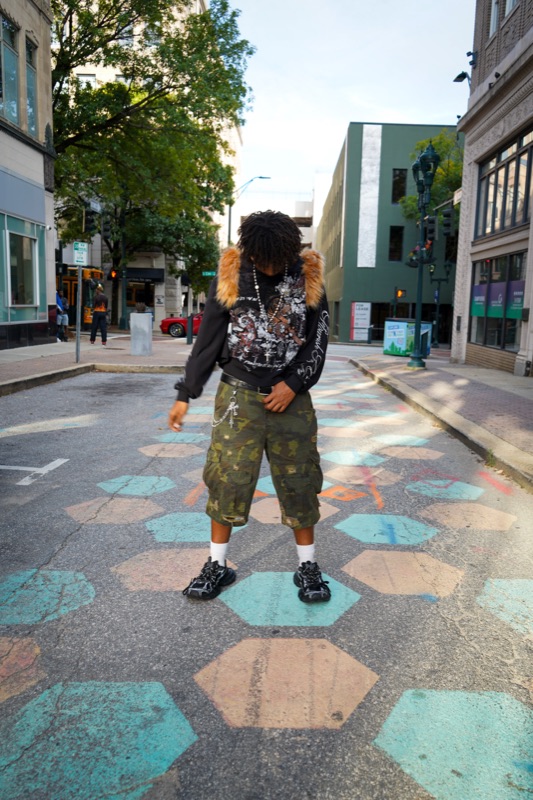 Young man in a fur-collared graphic sweatshirt looking down at painted hexagons on a downtown street.