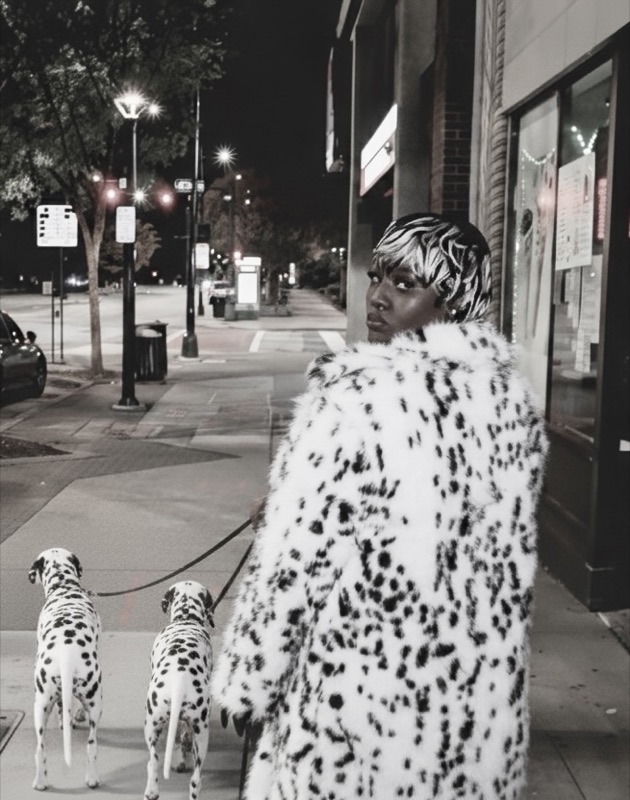 Black-and-white night scene of a woman in a dalmatian-print faux fur coat walking two dalmatians.