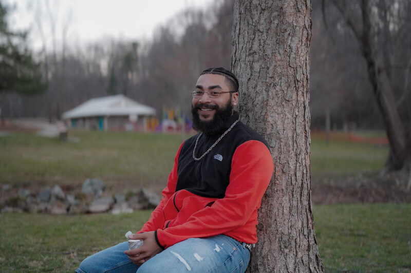 Smiling bearded man in a red-and-black North Face fleece leaning against a tree in a park at dusk.